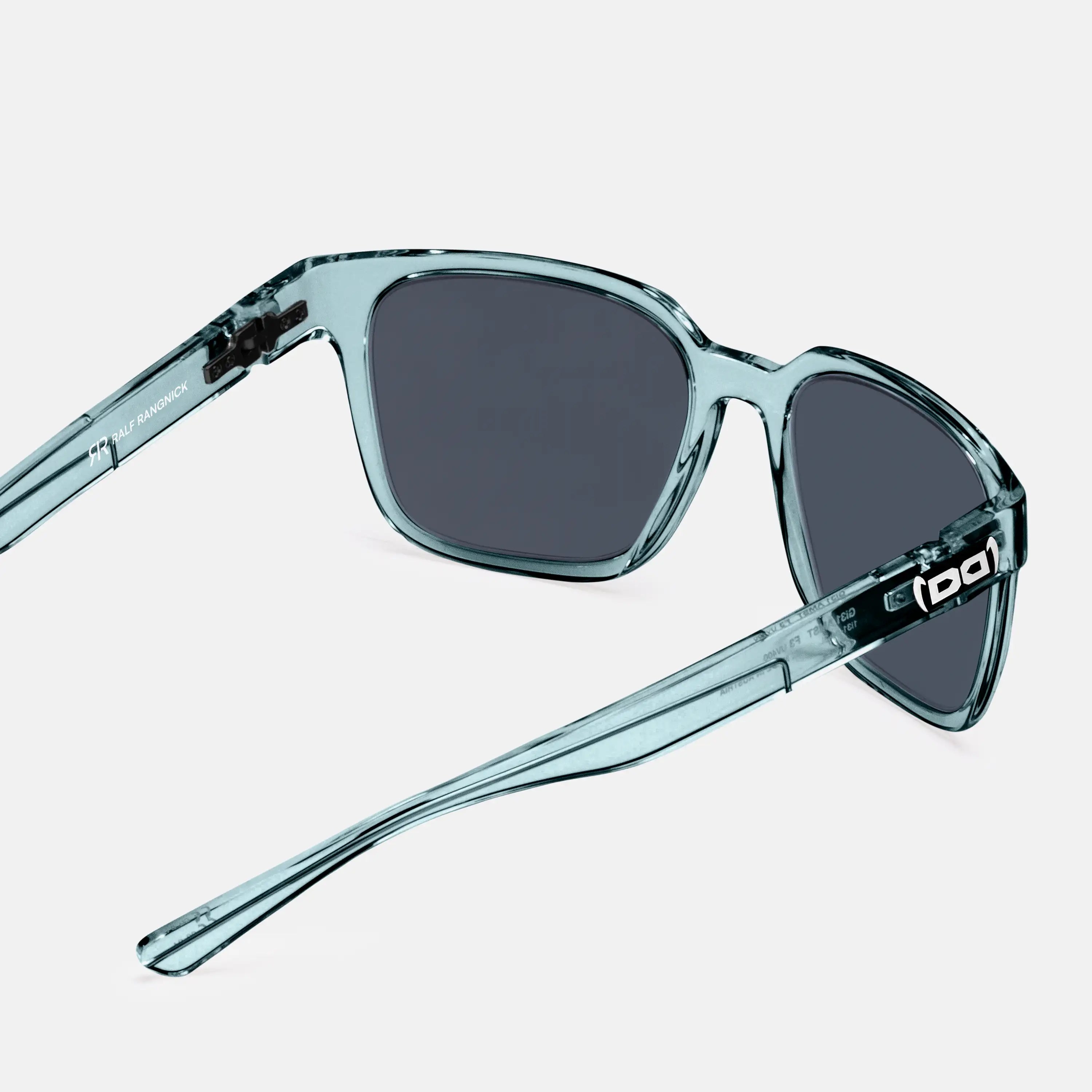 Eine stilvolle Sonnenbrille mit transparentem, hellblauem Rahmen und dunklen Gläsern liegt auf einer weißen Oberfläche. Die Bügel tragen dezente Markendetails, darunter der Schriftzug „Ralf Rangnick“ auf der Innenseite. Das Design ist modern und minimalistisch.
