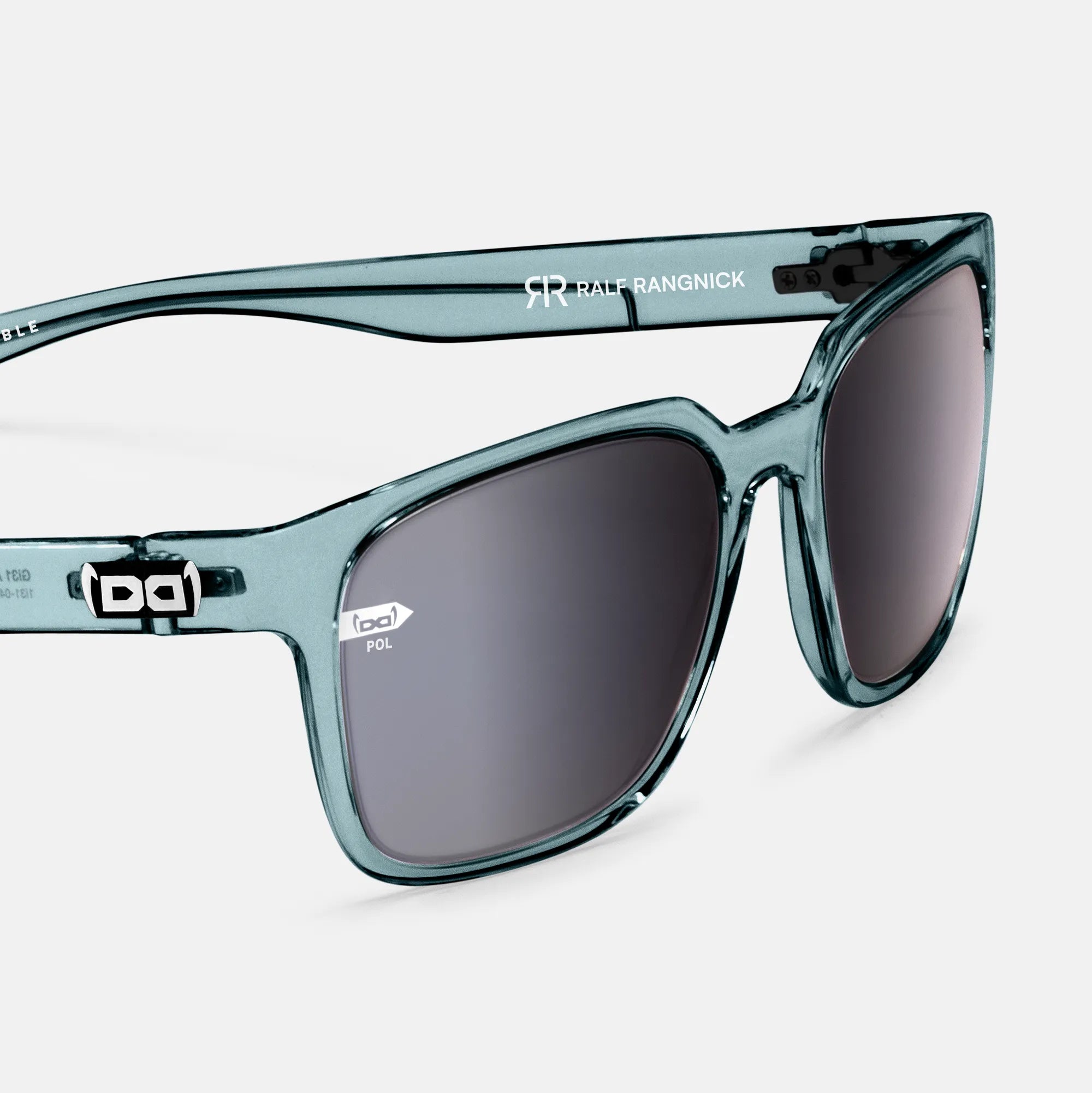 Eine moderne Sonnenbrille mit transparentem, hellblauem Rahmen und polarisierten Gläsern in einem dunklen Farbton. Der Bügel trägt das Logo „RR Ralf Rangnick“ sowie das Markenlogo „bd“. Das Design kombiniert Eleganz mit sportlichem Stil.