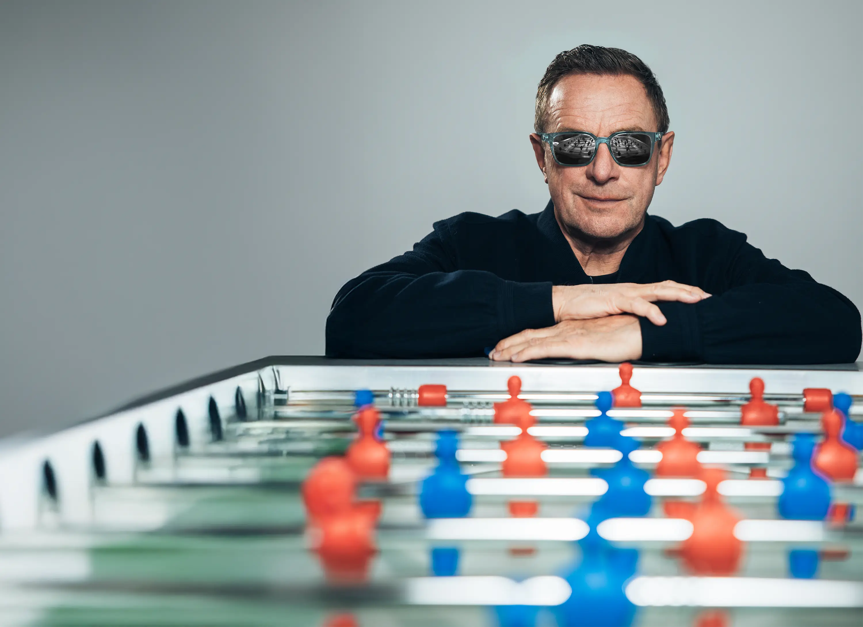 Ein Mann mit kurzen Haaren und dunkler Jacke lehnt entspannt an einem Tischkicker. Er trägt eine Sonnenbrille mit verspiegelten Gläsern, in denen sich der Tischkicker spiegelt. Der Fokus liegt auf seinem selbstbewussten Ausdruck, während die Spielfiguren des Tischkickers im Vordergrund leicht unscharf sind.
